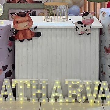 A close-up of the personalized details for Atharva's farm-themed party. This shot shows the light-up marquee letters of his name and cute animal cutouts on the dessert table.