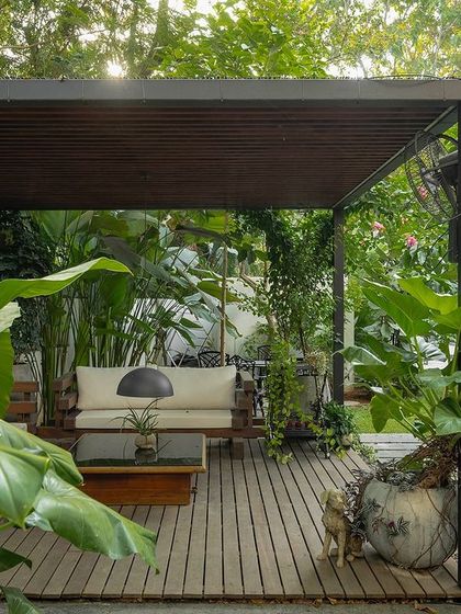 A covered deck with comfortable seating, nestled within a lush garden. This outdoor living room is framed by large plants and a pergola, offering a perfect blend of comfort and nature.
