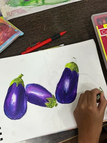 A student uses oil pastels to draw brinjals, learning blending techniques to show highlights and shadows. We teach various mediums as required by the elementary and intermediate exam curriculum.