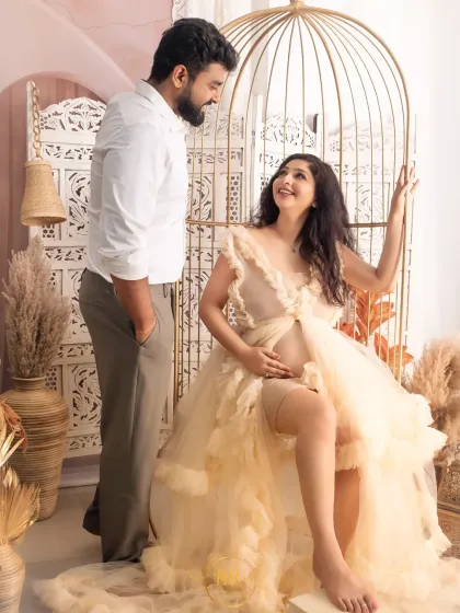 A sweet interaction between partners in a bohemian-themed setup. The mother-to-be is seated in a golden birdcage prop while her partner looks on lovingly.