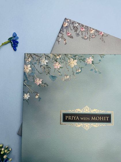 A close-up of a pastel blue invitation adorned with delicate, embossed white and pink flowers, creating a soft and romantic feel.