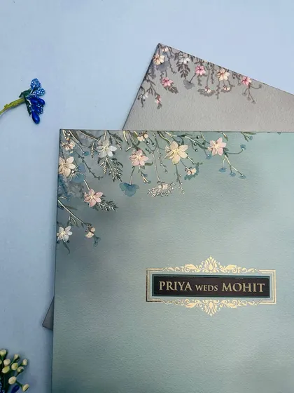 A close-up of a pastel blue invitation adorned with delicate, embossed white and pink flowers, creating a soft and romantic feel.