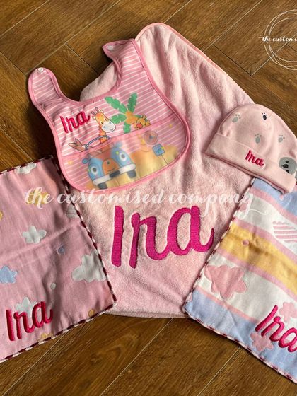 A complete newborn baby set in pretty pink for baby Ira. This collection includes a large personalized towel, custom-embroidered napkins, a bib, and a matching cap for a coordinated and thoughtful gift.