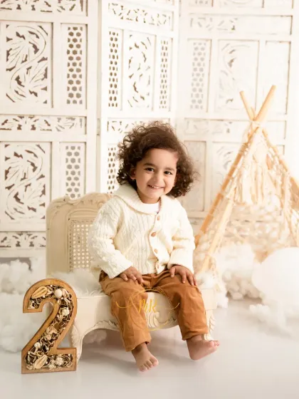 A happy two-year-old boy sits on a miniature bench next to a decorative number "2" in a bright, neutral-toned set.