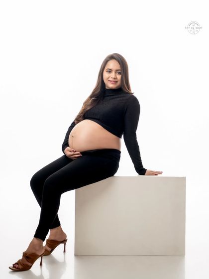 A chic and modern solo portrait of a mom-to-be in a stylish all-black outfit, seated against a clean white background.