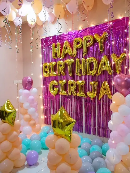 This birthday setup uses pastel-colored balloon pillars and loose balloons on the floor. A pink shimmer curtain with large 'Happy Birthday' foil letters serves as the main backdrop.