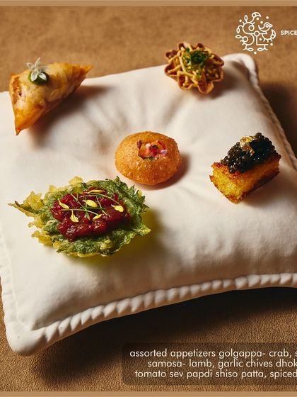 A whimsical presentation of our assorted appetizers, including lamb samosa and dhokla with caviar, served on a plush white pillow. It’s a statement of our playful yet sophisticated style.