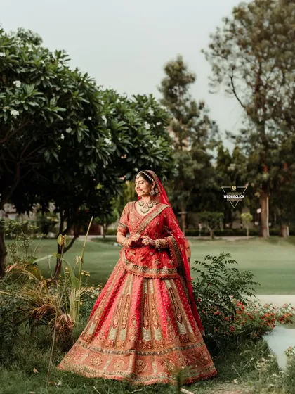 A full-length portrait of the bride in a garden setting, her traditional red and gold lehenga looking stunning against the natural greenery.