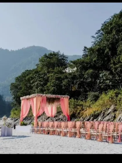 A wide shot of a destination wedding setup on a riverbank. The pink-draped mandap and guest seating look beautiful against the backdrop of the mountains and river.