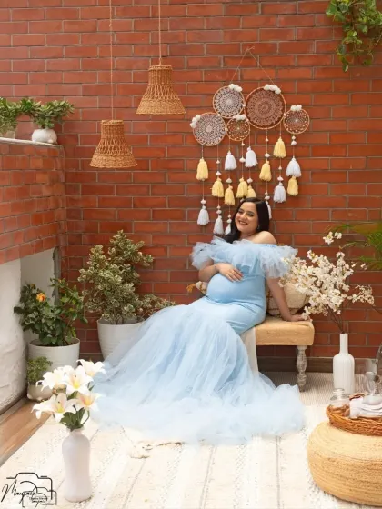 The full view of the cozy corner setup, with its brick wall, dreamcatchers, and lush plants. The mama-to-be looks serene in her flowing blue gown.