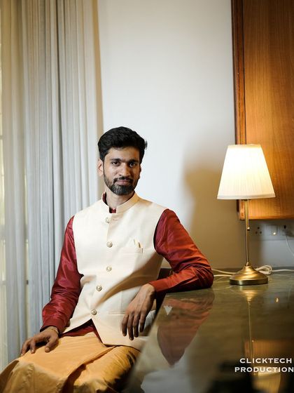 A dapper portrait of a groom in a traditional silk kurta and veshti with a modern waistcoat, seated in a well-lit, contemporary interior space.