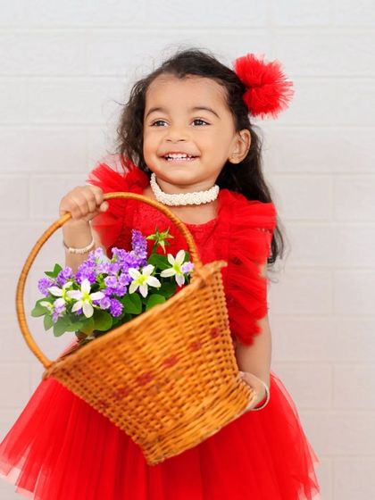 A smiling girl in a red dress holds a basket of flowers against a simple white brick wall. This classic and cheerful portrait is timeless and beautiful.