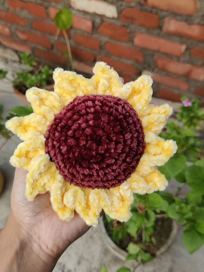 Holding the plush, soft-yarn sunflower outdoors against a brick and plant background.