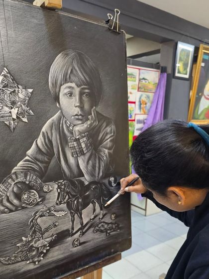 An over-the-shoulder view of the artist at her easel, working on the composition of her glass marking pencil drawing of a boy and his toys.