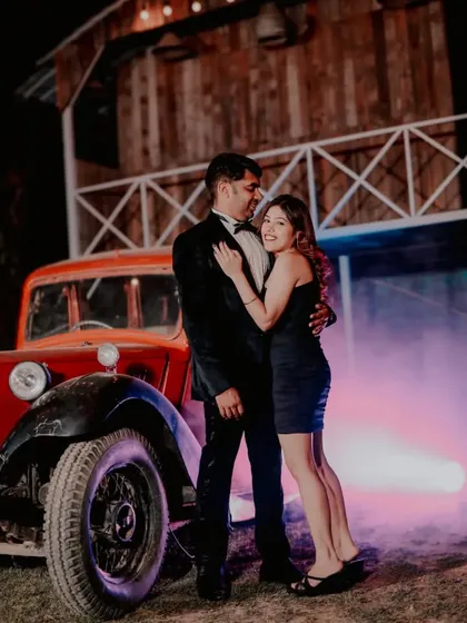 A stylish couple poses next to a vintage red car, with colored smoke adding a touch of modern drama to the rustic night setting. This showcases our creative use of props and effects for a unique pre-wedding photoshoot.