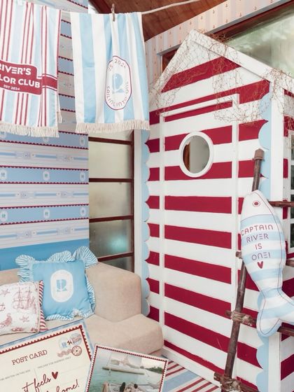 Welcome to 'River's Sailor Club,' a nautical-themed first birthday. This cozy corner features a custom-built red and white striped cabin, personalized pillows, and themed postcards.