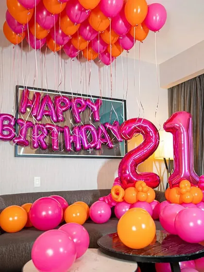 A vibrant 21st birthday setup in hot pink and orange, with balloons on the ceiling and a large number display.