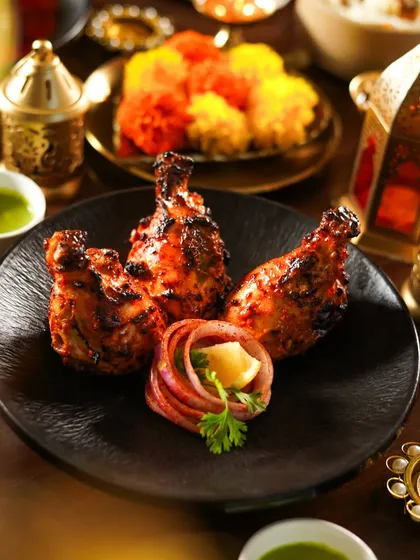 Tandoori chicken drumsticks arranged beautifully on a black plate with onion rings. The warm, festive lighting and colorful background make this a standout dish for any celebration.