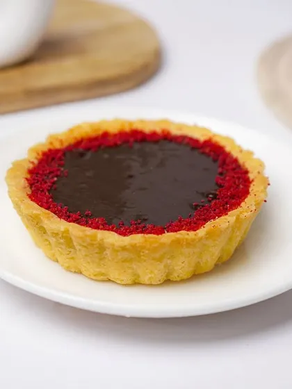 A red velvet chocolate tart with a glossy finish, presented on a simple white plate. The vibrant red and dark chocolate create a striking contrast in this clean product shot.