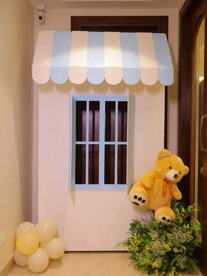 A charming prop designed for a baby shower, featuring a miniature house front with a blue and white striped awning and a cute teddy bear peeking out.