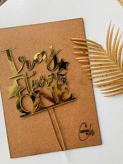 The "Iraj Turns ONE" topper in gold acrylic, presented on its backing board to showcase the quality and craftsmanship.