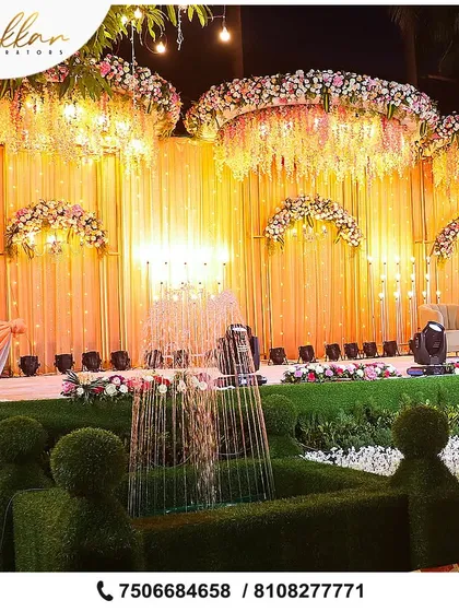 A grand reception stage with a unique fountain installation made of greenery. The backdrop of hanging flowers and warm lighting adds to the magical, garden-like feel.