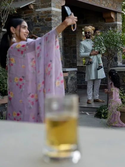 A creatively framed candid shot of a guest taking a selfie, with the groom and a child visible in the background.