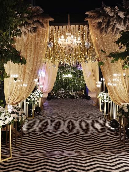 A grand entrance for a sangeet night, featuring a dramatic walkway with golden sequin drapes, crystal chandeliers, and a custom welcome sign for the couple.