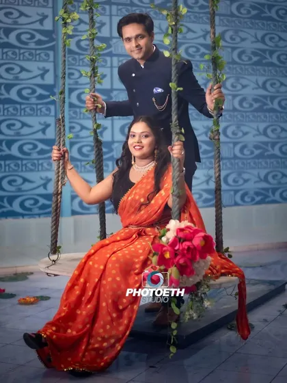 A sweet and traditional indoor pre-wedding photo on a decorated swing. The bride's orange saree adds a beautiful pop of color to the scene.