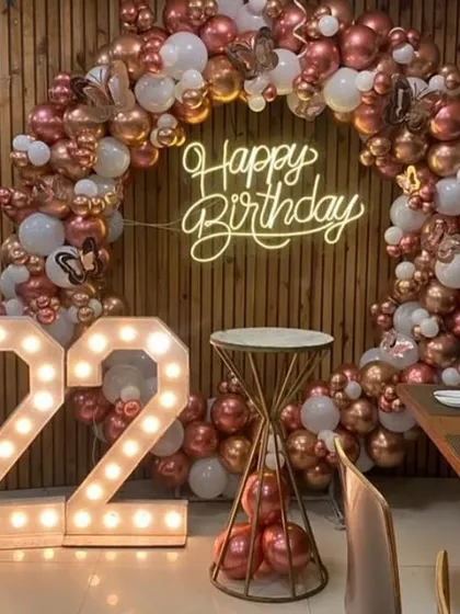 A chic and modern setup for a 22nd birthday celebration. The focal point is a large balloon ring in rose gold and white, accented with butterfly details and a custom 'Happy Birthday' neon sign, complemented by large marquee numbers.