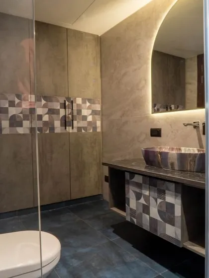 This bathroom showcases a playful use of geometric tiles on the vanity and as a decorative band in the shower. The backlit arched mirror and unique vessel sink add to its contemporary character.