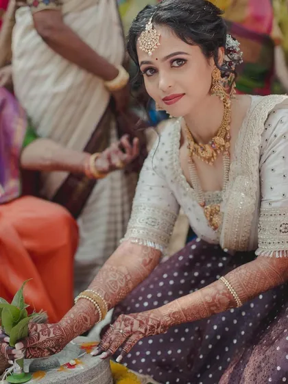 A bride participating in a wedding ritual, wearing a custom off-white blouse with a purple polka-dot lehenga skirt.