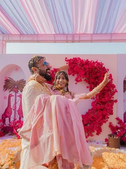 A playful moment at the end of a Haldi ceremony, with the groom carrying the bride against a custom-designed backdrop featuring pink giraffes and bougainvillea.