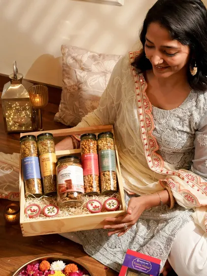 A woman in traditional attire admiring a Diwali gift hamper filled with healthy munchies. The warm lighting from the lantern creates a cozy, festive glow.