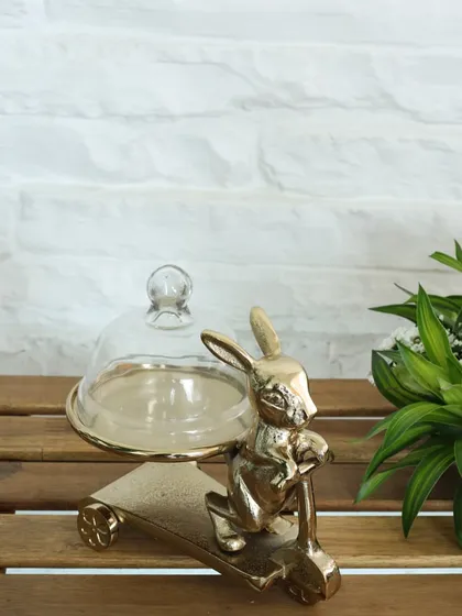 A touch of whimsy for your table. This golden stand features a rabbit on a scooter, holding a small platter with a glass cloche, perfect for serving a special treat or as a decorative accent.