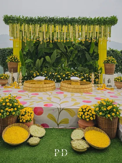 A full view of a traditional Haldi stage, decorated with marigolds, banana leaves, and printed fabric. The setup is vibrant, auspicious, and picture-perfect.
