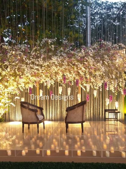 An ethereal stage perfect for photos. The backdrop of thin vertical slats is adorned with a cascade of white and purple flowers, while the clear stage is lit from below by dozens of candles.