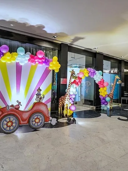An exciting indoor carnival setup featuring a vibrant photo booth with a circus car cutout. The scene is brought to life with a colorful balloon arch, life-sized animal props like a zebra and giraffe, and a multi-activity cart.