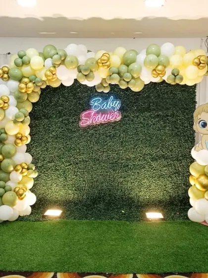 A simple and elegant baby shower photo booth. This design uses a green hedge wall as the backdrop, a 'Baby Shower' neon sign, and a balloon arch in muted green, white, and gold tones.