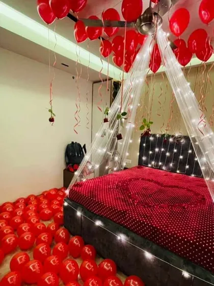A classic romantic room decoration featuring a bed canopy with fairy lights, red balloons covering the floor and ceiling, and a heart made of rose petals on the bed.