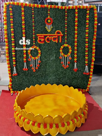 A clear view of our Haldi decor package. This shows the artificial grass wall with marigold borders, hanging floral elements, the 'हळद' sign, and the large golden Urli ready for the ceremony.