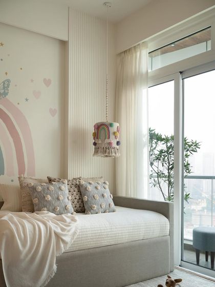 A custom pendant light hangs over the grey sofa bed, adding a whimsical touch to this daughter's bedroom. The pastel rainbow mural and soft textures create a magical and comforting atmosphere.