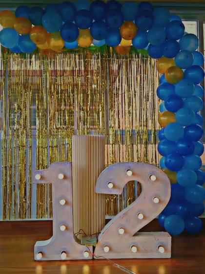 A simple and clean setup for a 12th birthday. This decoration features large, illuminated '12' marquee numbers and a blue and yellow balloon arch against a gold tinsel curtain, perfect for at-home celebrations.