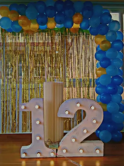 A simple and clean setup for a 12th birthday. This decoration features large, illuminated '12' marquee numbers and a blue and yellow balloon arch against a gold tinsel curtain, perfect for at-home celebrations.