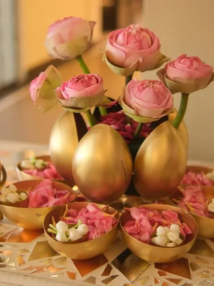 It's the little details that make an event truly special. This centerpiece features delicate pink lotus buds in golden egg-shaped vases, surrounded by flower petals and a small golden elephant, adding a unique and elegant touch to the table decor.