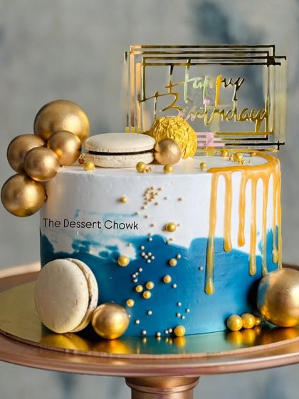A modern drip cake in blue and white, decorated with gold macarons, spheres, and a stylish topper. The golden drip adds a touch of luxury.