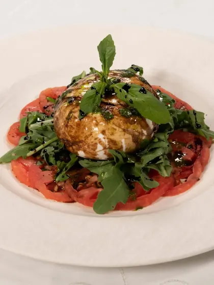 A fresh Burrata salad, served on a bed of sliced tomatoes with rocket leaves and a balsamic glaze. It is a creamy, fresh, and delicious starter.