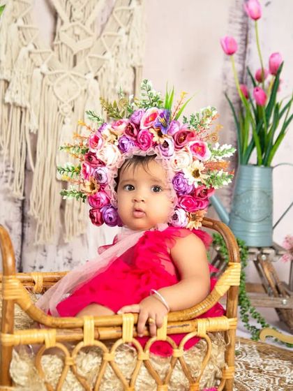 A lovely portrait from a half-birthday floral photoshoot. The baby is sitting in a wicker pram, surrounded by pink flowers and wearing an elaborate floral headpiece.