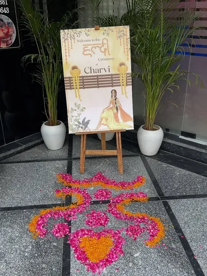 A personalized welcome area for a Haldi ceremony, featuring a custom welcome sign on an easel and a beautiful flower rangoli on the floor.