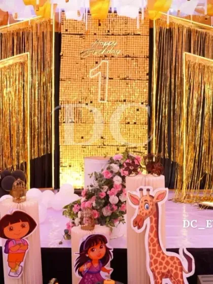 A fun-filled first birthday stage with a 'Dora the Explorer' theme. The gold fringe backdrop, character cutouts, and balloon arrangements create an exciting and adventurous atmosphere for the party.